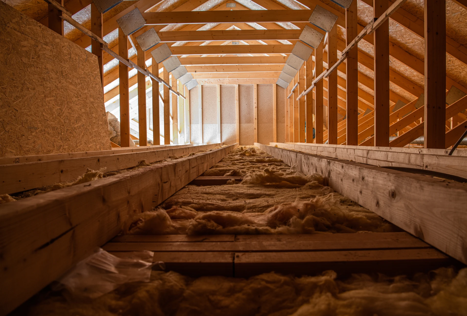 Vue intérieure d'un comble en cours d'isolation, montrant des panneaux d'isolant thermique (laine minérale, ouate ou fibre de bois) en train d'être posés entre les chevrons d'une charpente en bois. L'artisan est visible en train d'installer ou d'ajuster l'isolant. La lumière naturelle provient des fenêtres de toit (Velux). L'ambiance est claire et professionnelle, mettant en avant le matériau isolant et le travail de couverture artisanal. Comble de maison individuelle, contexte résidentiel moderne.