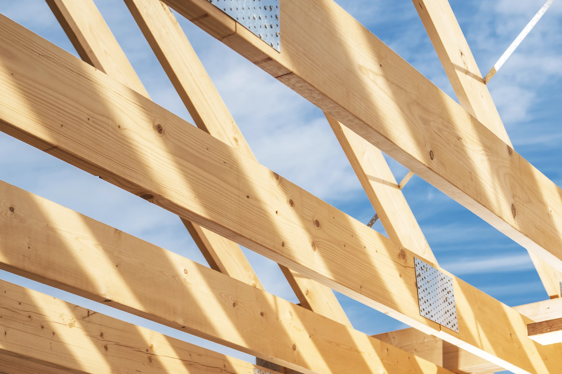 Charpente bois neuve en cours de pose sur chantier de rénovation résidentiel, vue en plongée partielle. Poutres principales et secondaires en bois massif clair fraîchement équarries, assemblages traditionnels visibles à mi-bois ou par boulons galvanisés. Sous-toiture grise HPV déroulée sur les chevrons, liteaux et contre-liteaux en bois jaune disposés en grille régulière. Chantier en cours dans contexte résidentiel breton ou français, architecture traditionnelle visible en arrière-plan, ciel dégagé, éclairage naturel mettant en avant la solidité et la qualité du travail.